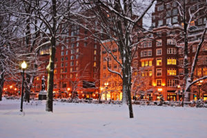 Snow-covered park with city buildings lit warmly at dusk.